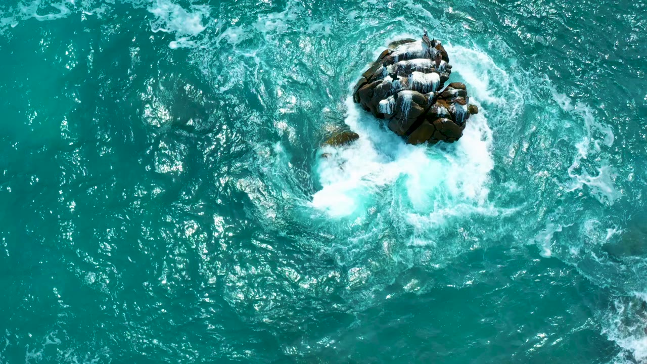 Overhead view at Secrets Marquis Los Cabos, Mexico, as a brighter burst of white surf pushes into the lower side of the rock cluster in clear turquoise water, with no visible people.