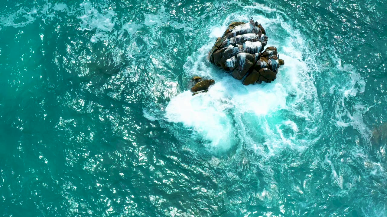 Direct overhead frame at Secrets Marquis Los Cabos, Mexico, with a broad white foam patch spreading below the main rock cluster and smaller rocks beginning to show through the water, with no visible people.