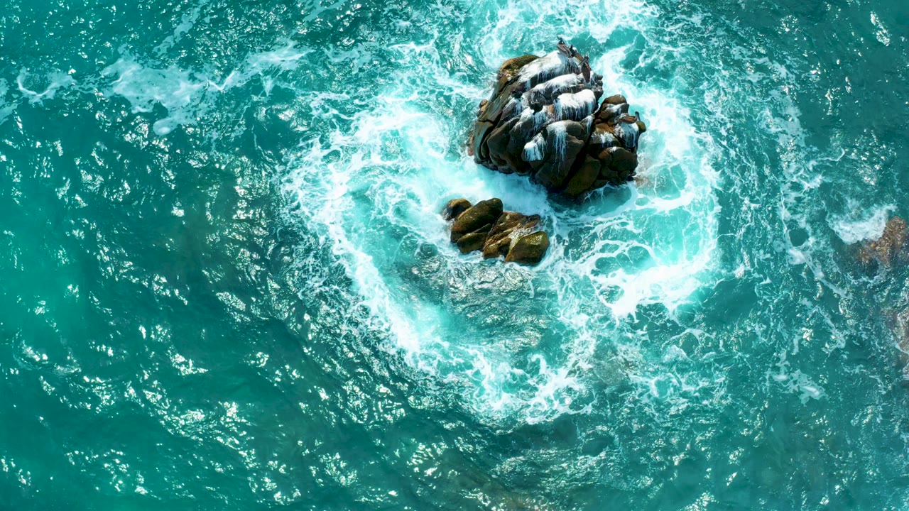 Overhead view at Secrets Marquis Los Cabos in Mexico shows turquoise water and white surf circling a central dark rock cluster, with no visible people.