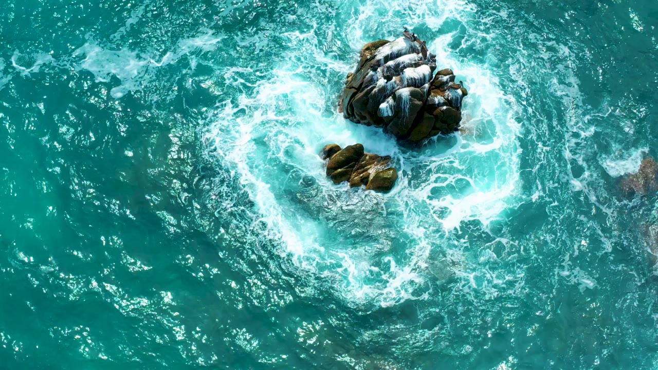 A top-down frame from Secrets Marquis Los Cabos in Mexico shows white foam wrapping more tightly around the central rocks as teal water fills the surrounding frame, with no visible people.