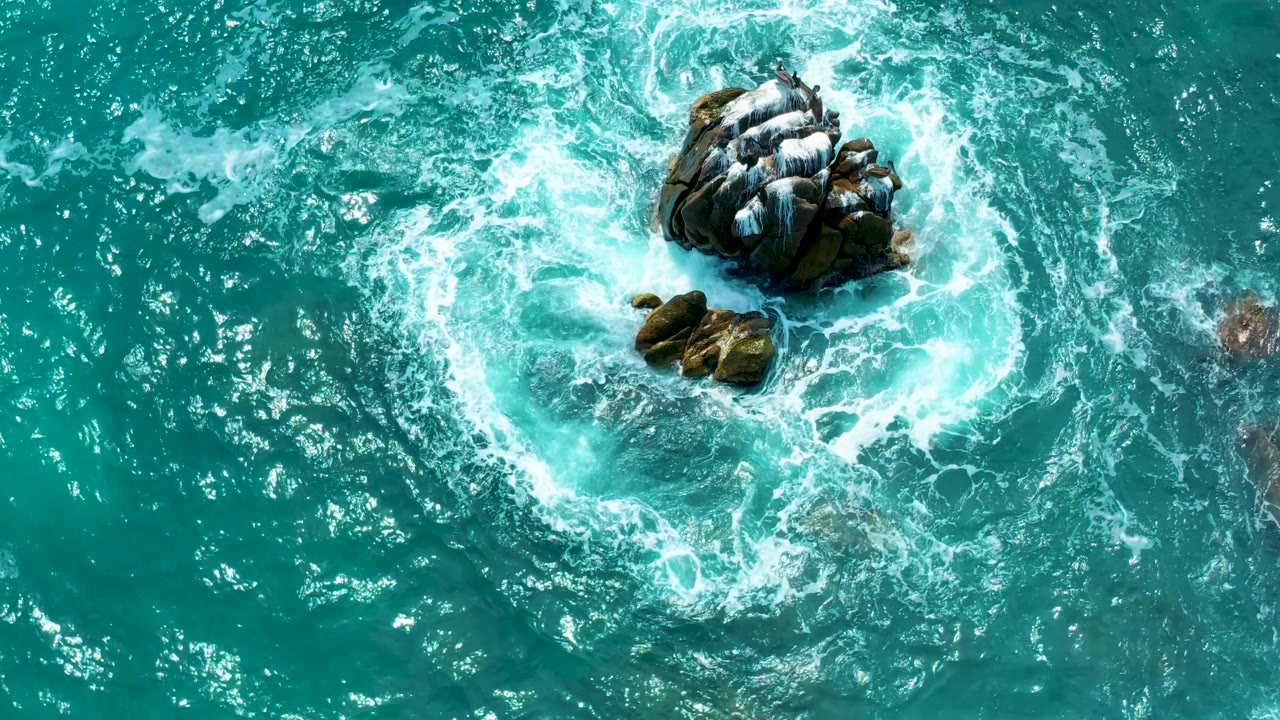 The Mexico coastal aerial shows the main rock cluster near the upper center with white wash spreading across clear green-blue water and smaller rocks below, with no visible people.
