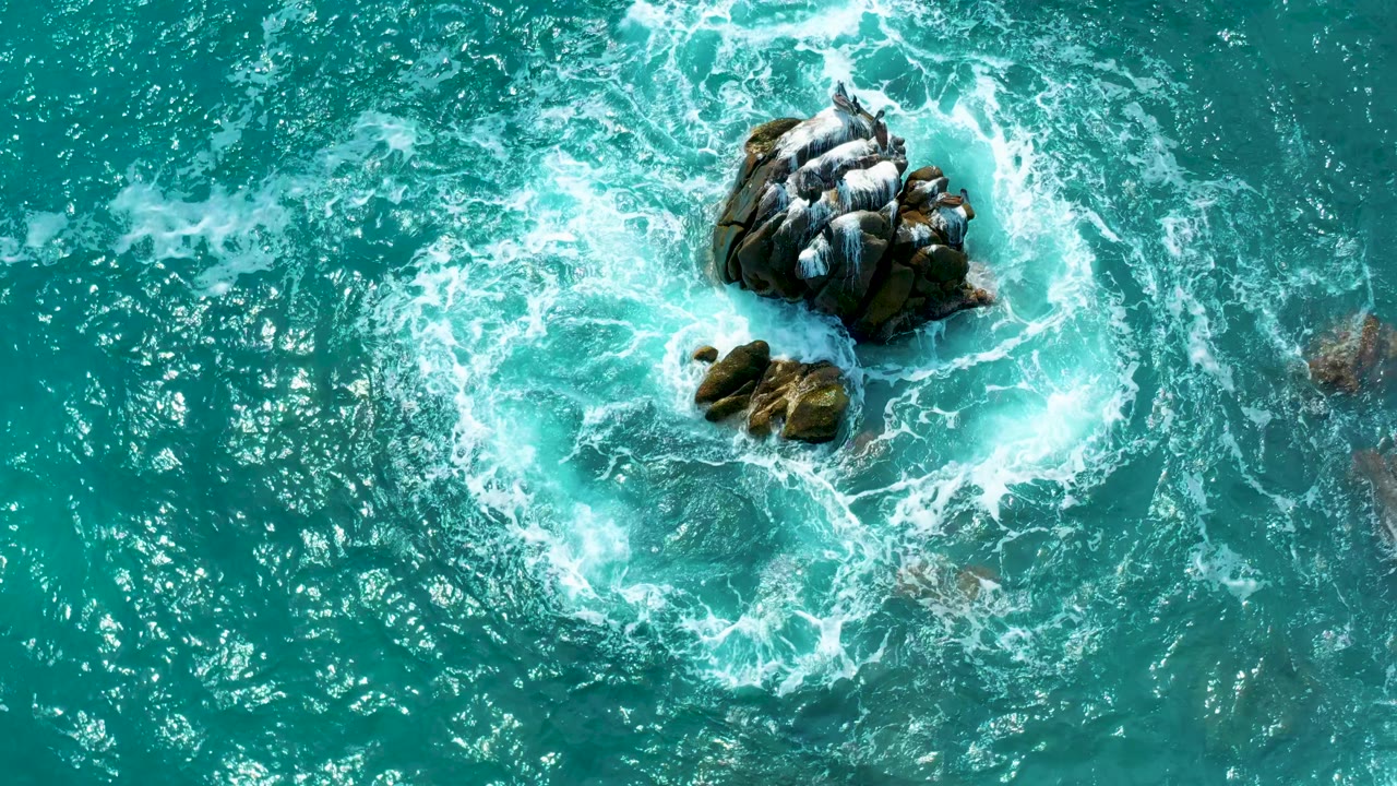 An overhead Los Cabos frame shows the large dark rocks shifted toward the upper part of the image, with small stones below and foamy surf breaking around both, with no visible people.