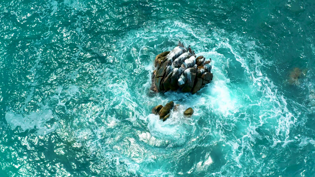 Overhead view at Secrets Marquis Los Cabos in Mexico showing turquoise water and white surf wrapping around a central rock cluster, with no visible people.