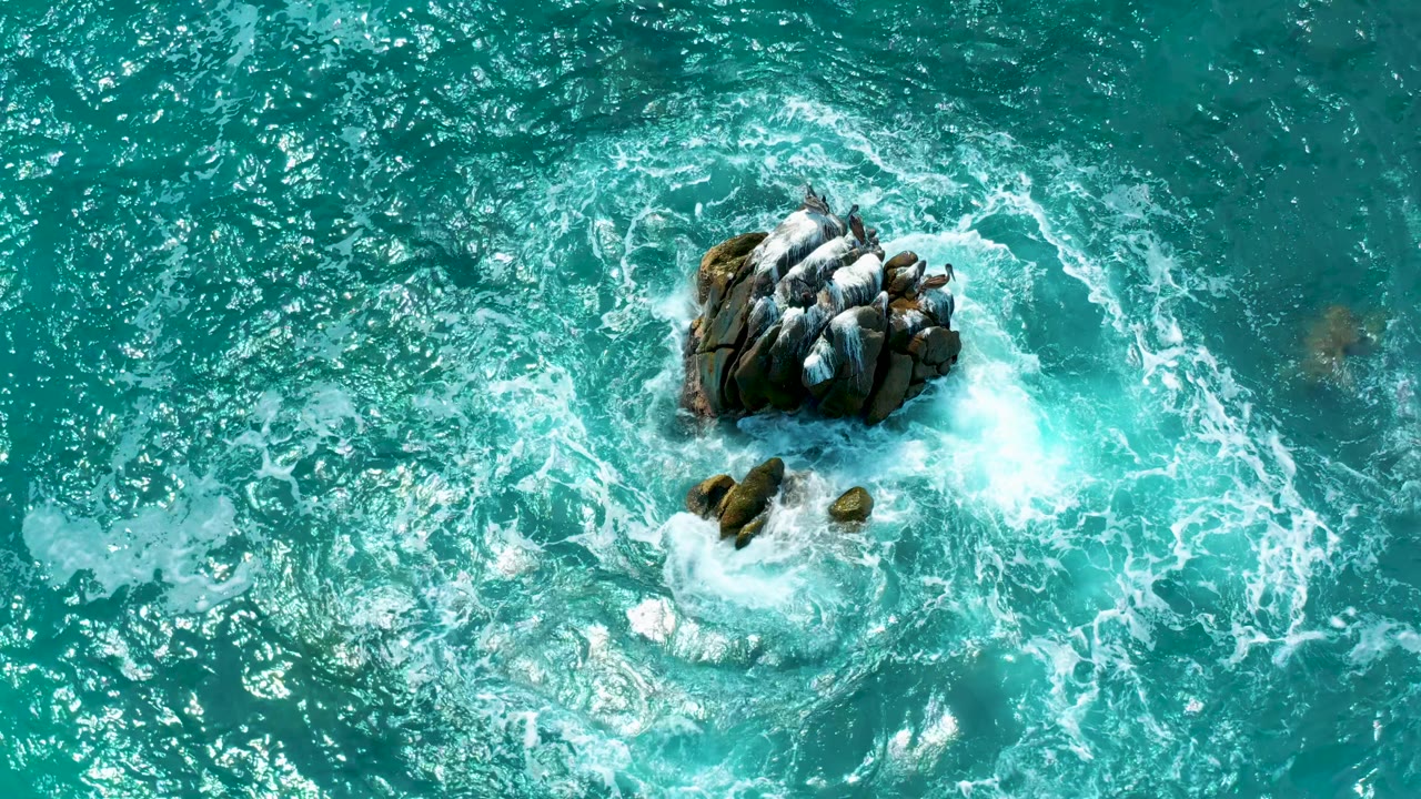Aerial frame from Secrets Marquis Los Cabos in Mexico with bright foam pushing along the right side of the main rocks and darker submerged shapes nearby, with no visible people.