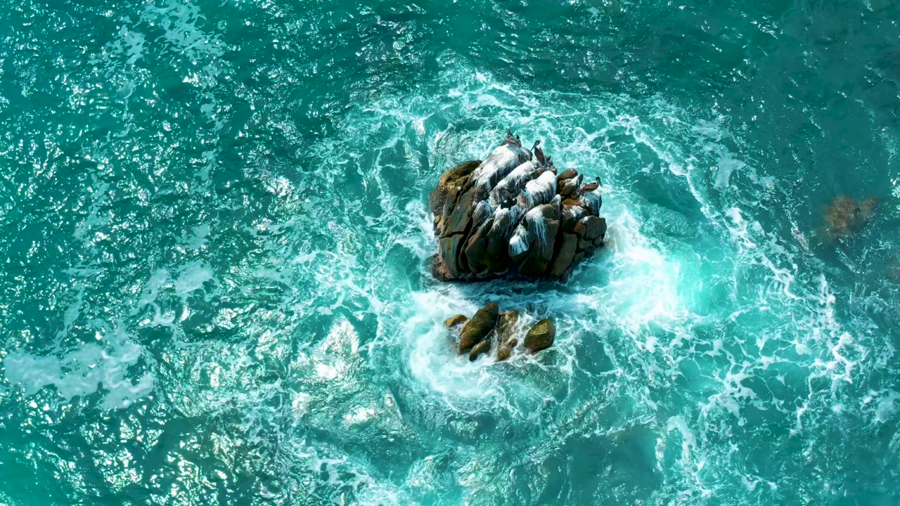Top-down view at Secrets Marquis Los Cabos in Mexico where the main rock outcrop sits high in frame as foam curls around smaller rocks below, with no visible people.