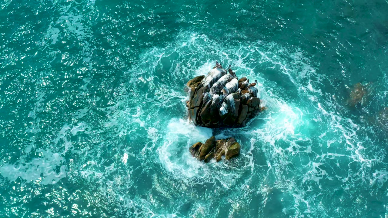 Drone-like overhead view from Secrets Marquis Los Cabos in Mexico showing waves circling the left and lower edges of a rocky formation, with no visible people.