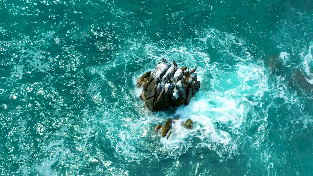 Turquoise waves swirl around an isolated rock outcrop.
