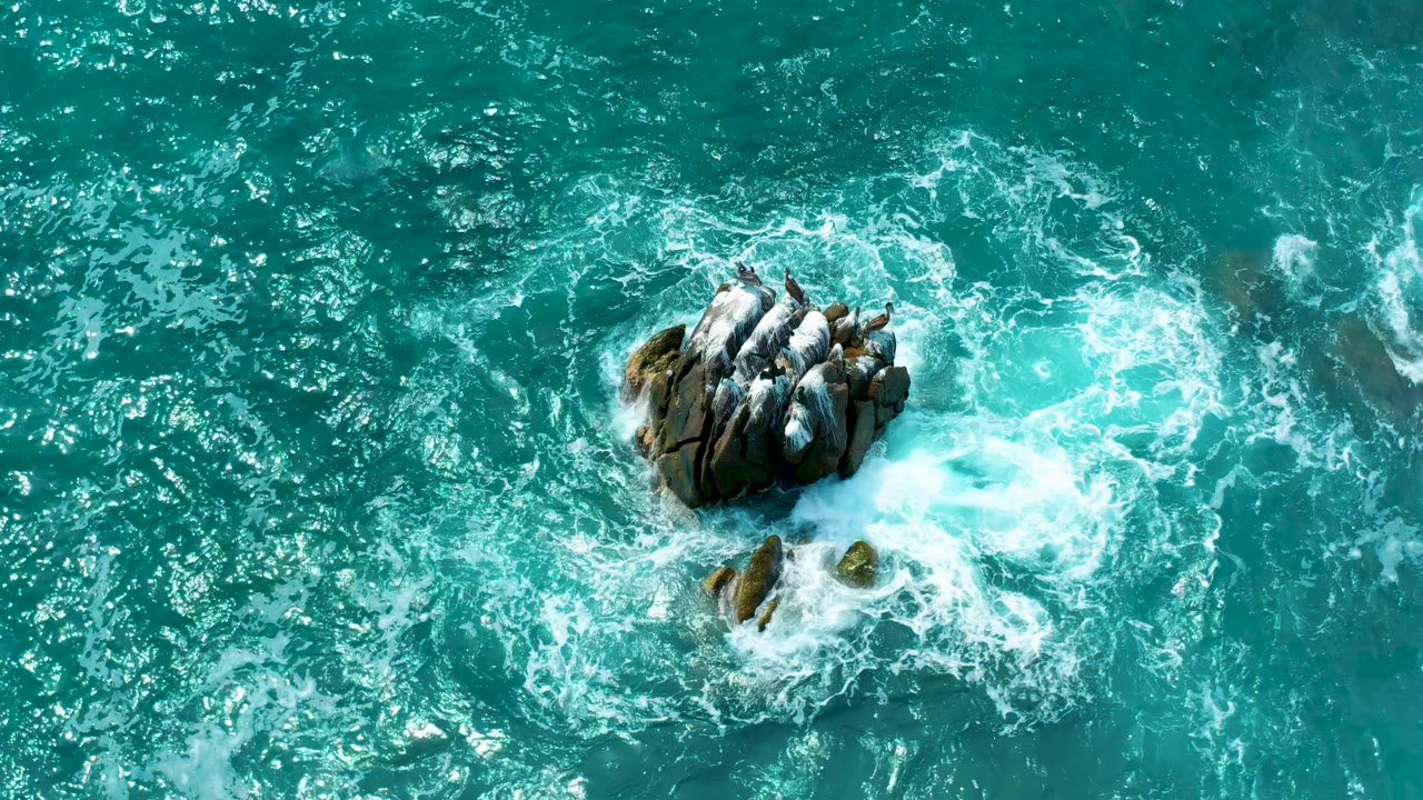 Aerial frame of foamy water tightening around the rugged rock as darker submerged shapes show beneath the teal sea at Secrets Marquis Los Cabos, Mexico, with no visible people.
