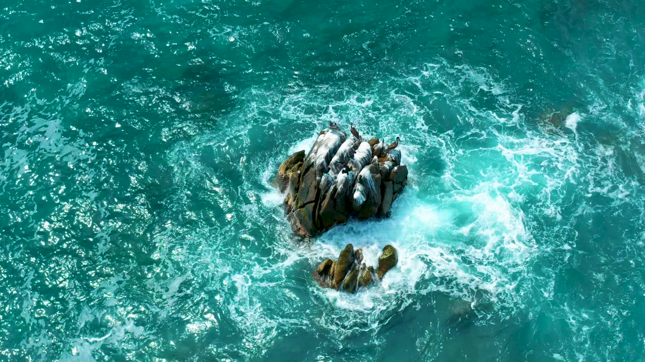 Overhead view of the rock shifted slightly toward the upper right, with foam trails spreading across turquoise water at Secrets Marquis Los Cabos, Mexico, and no visible people.