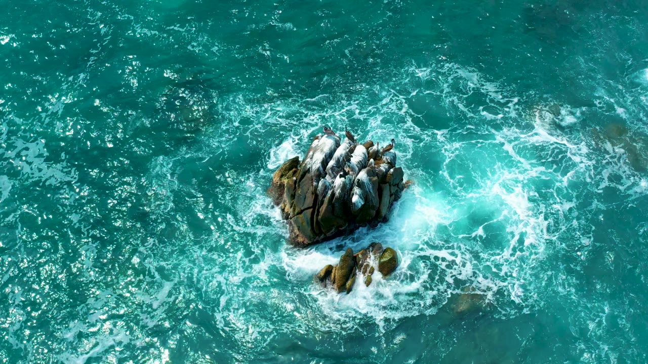 Aerial view of white surf breaking around the main rock and a smaller rock cluster below it in bright turquoise water at Secrets Marquis Los Cabos, Mexico, with no visible people.