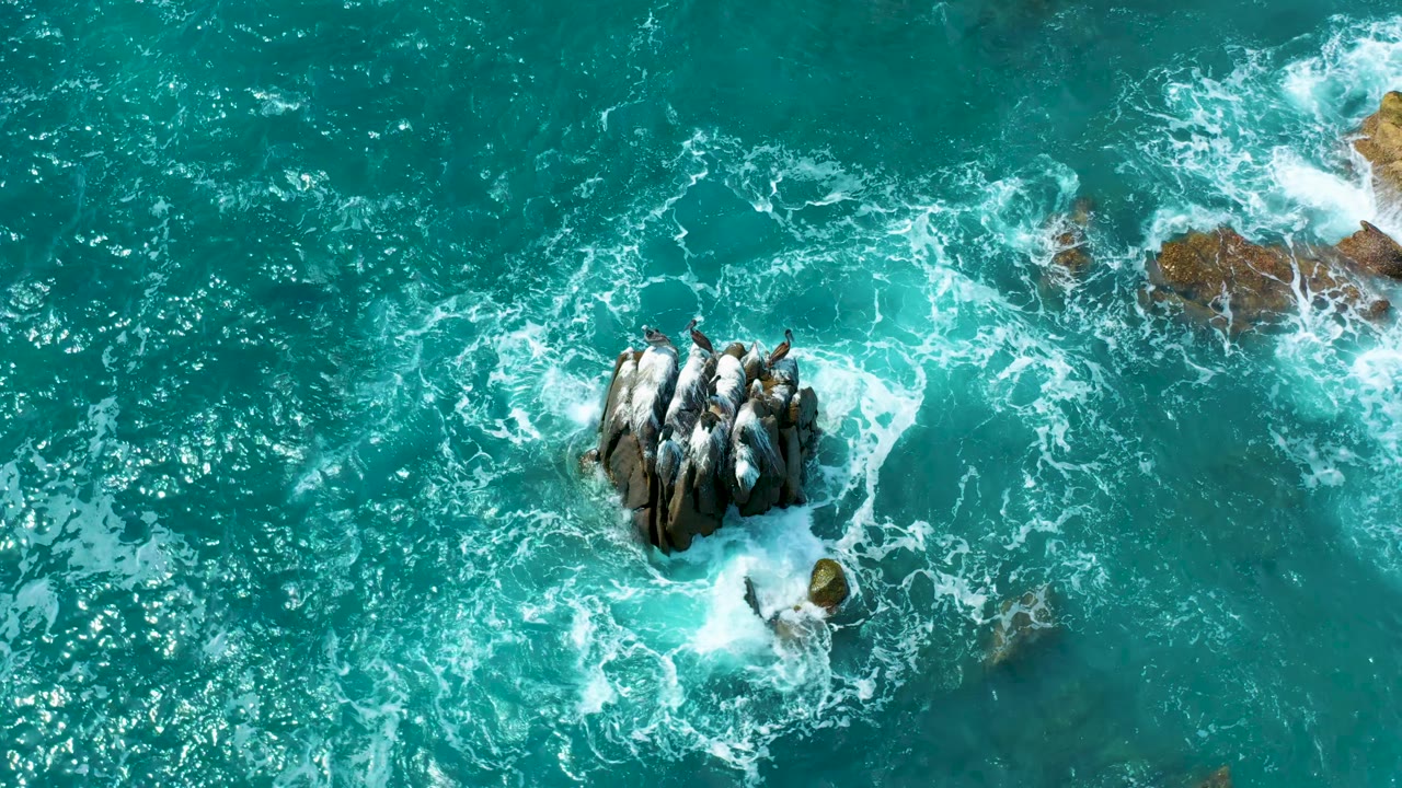 Aerial frame at Secrets Marquis Los Cabos, Mexico, with the dark rock cluster centered as white foam spreads left and right across blue-green water, no visible people.
