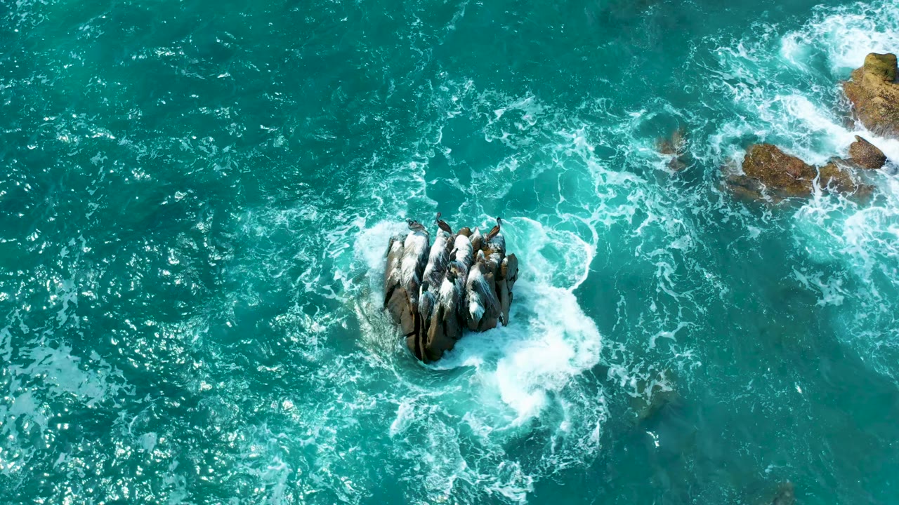 Top-down ocean view at Secrets Marquis Los Cabos, Mexico, where a compact rock cluster sits in turquoise surf with scattered white foam and no visible people.