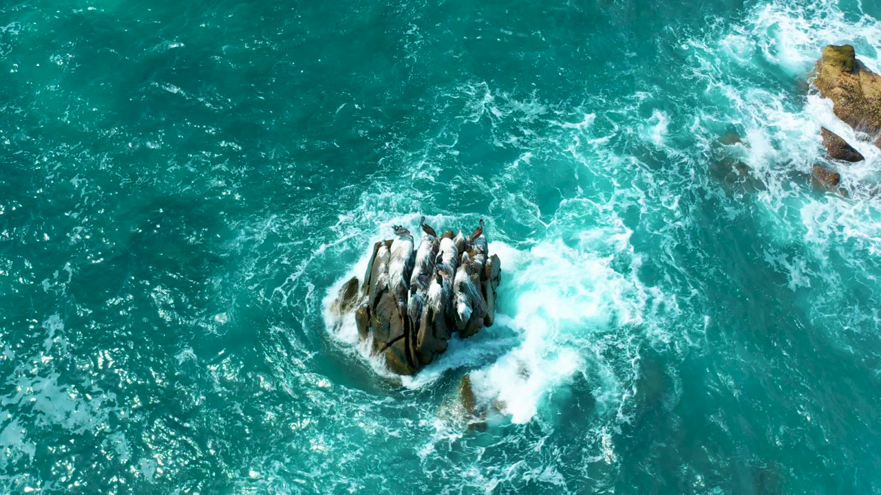 High aerial frame from Secrets Marquis Los Cabos, Mexico, showing teal water, white wave foam, a central rock cluster, and smaller rocks near the top edge with no visible people.