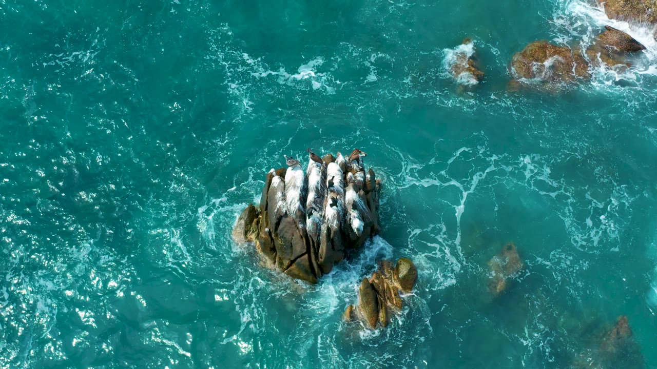 Overhead Los Cabos resort-coast water frame with the main rock cluster lower left, bright sun sparkle to the right, white foam around rocks, and no visible people.