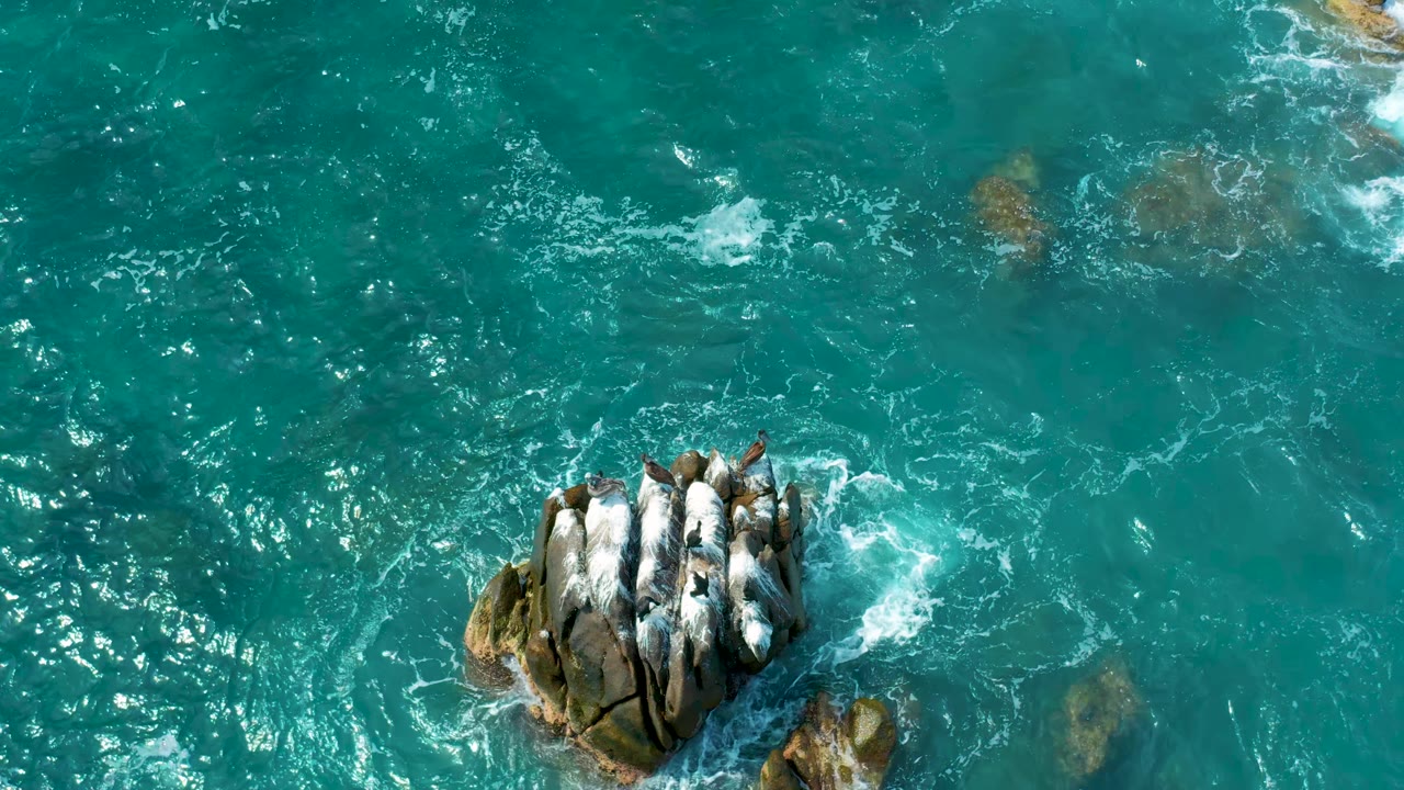 Aerial frame of blue-green water and a surf-washed rock outcrop at Secrets Marquis Los Cabos, Mexico, with foam concentrated on the right and no visible people.