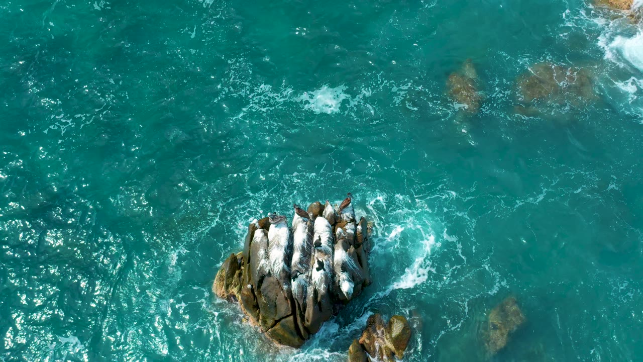 Overhead drone-style view of turquoise ocean at Secrets Marquis Los Cabos, Mexico, with the rock cluster lower in frame, bright water ripples to the left, and no visible people.
