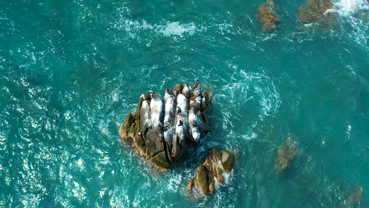 Aerial frame of blue-green sea breaking into white foam around tan rocks at Secrets Marquis Los Cabos, Mexico, with no visible people.