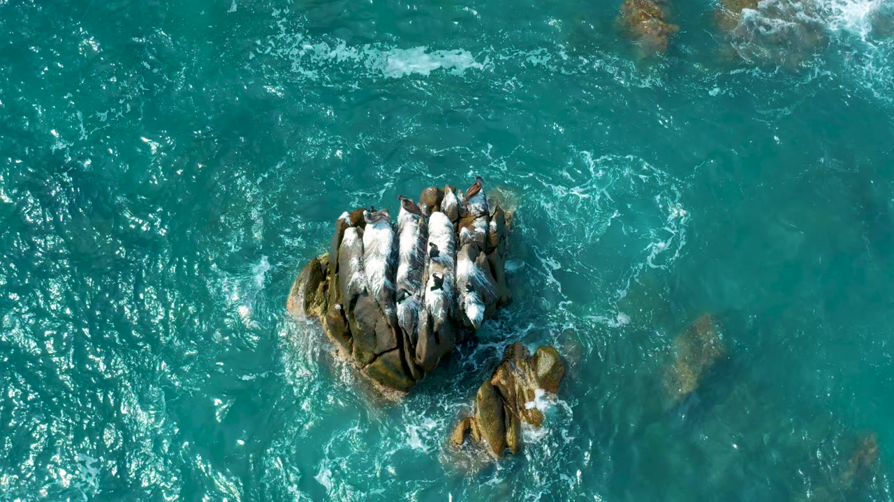 Top-down view of the rock cluster held near center as turquoise water and loose foam trails move around it at Secrets Marquis Los Cabos, Mexico, with no visible people.