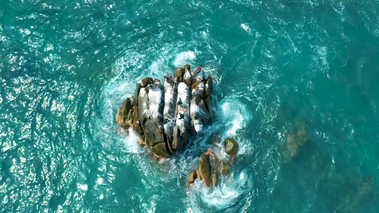 Aerial frame from Secrets Marquis Los Cabos, Mexico, with foam collecting on the upper edge of the main rocks and clear blue-green water around them, no visible people.