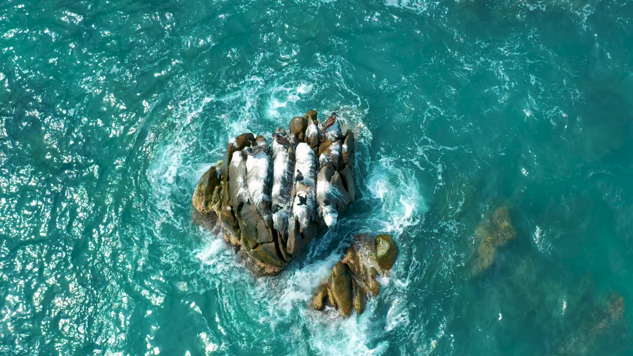 Straight-down coastal view at Secrets Marquis Los Cabos, Mexico, where rippled turquoise water surrounds the rock cluster and a smaller rock sits below it, no visible people.