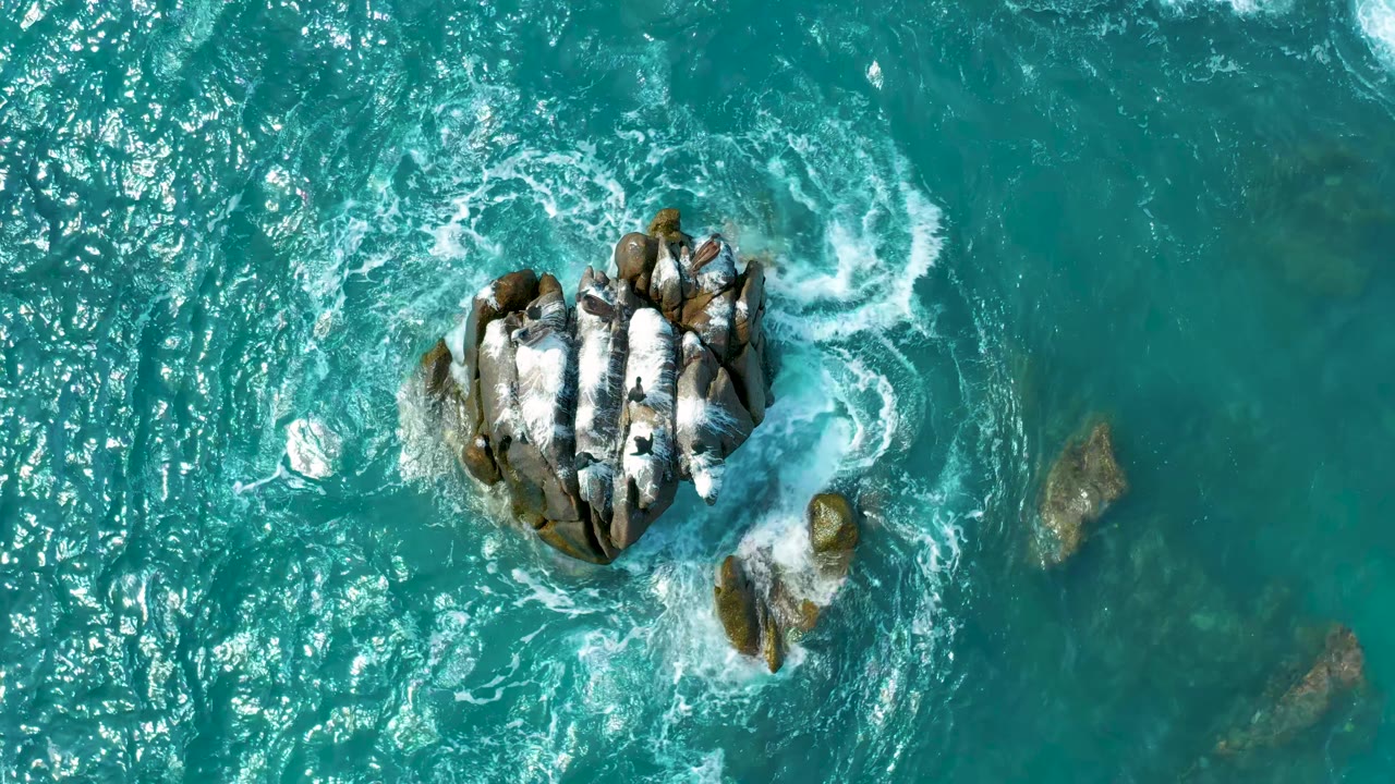 Overhead Los Cabos, Mexico view with the main rock cluster near center and a curved patch of white water wrapping around its upper-right edge, with no visible people.