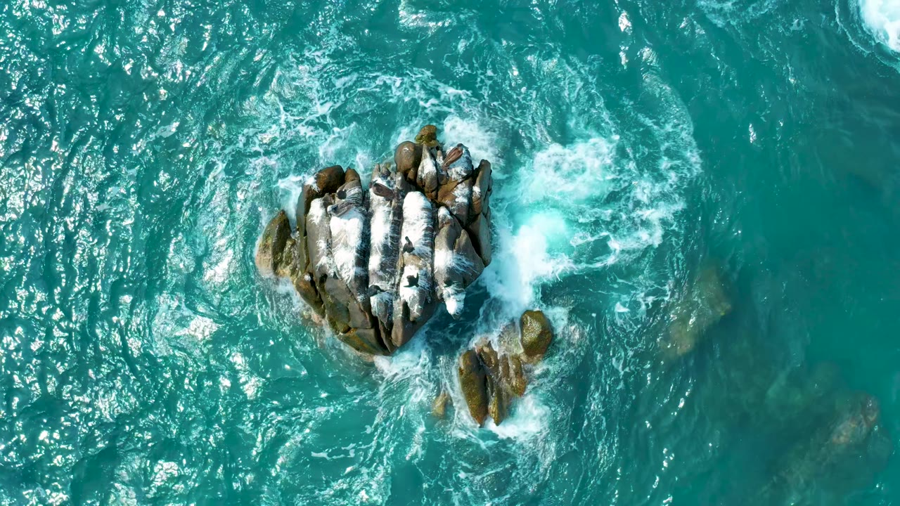 Overhead frame from Los Cabos, Mexico with white surf spreading away from the rock cluster and faint submerged rocks visible through the blue-green water, with no visible people.
