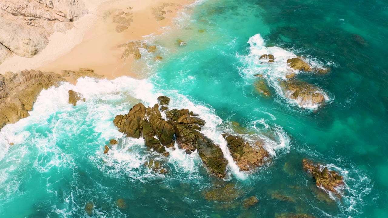 Overhead view at Secrets Marquis Los Cabos, Mexico, with turquoise water wrapping around dark coastal rocks, tan sand at the top, and no visible people.