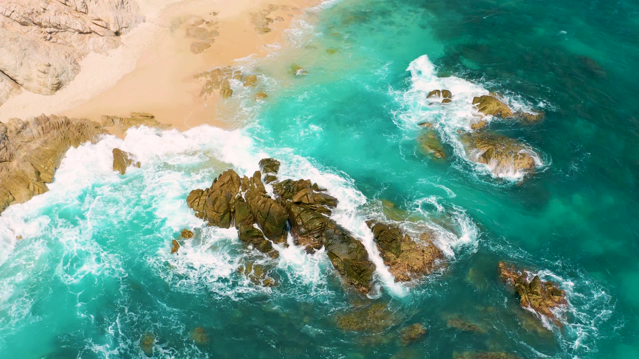Aerial frame at Secrets Marquis Los Cabos, Mexico, showing foamy waves pressing around a central brown rock formation with pale beach above and no visible people.
