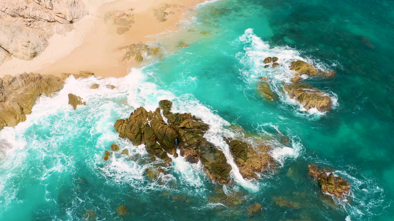 Drone-like overhead view at Secrets Marquis Los Cabos, Mexico, with the rock cluster shifted lower in frame, white surf curling near the beach, and no visible people.