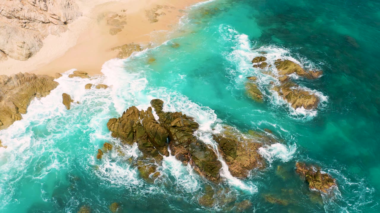 Overhead frame at Secrets Marquis Los Cabos, Mexico, with bright foam breaking around large brown rocks, beach and cliff textures at the top, and no visible people.