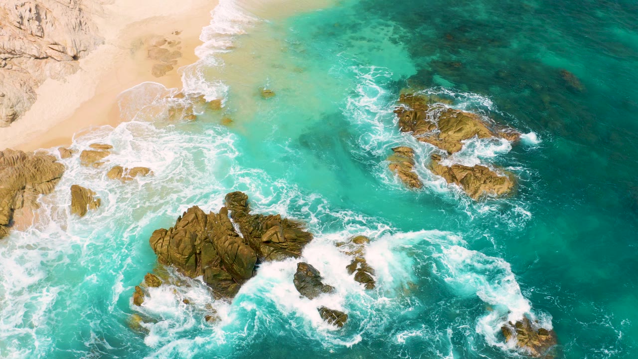 Aerial frame over the Mexico shoreline with pale sand at upper left, rock outcrops in the surf, turquoise water across the frame, and no visible people.