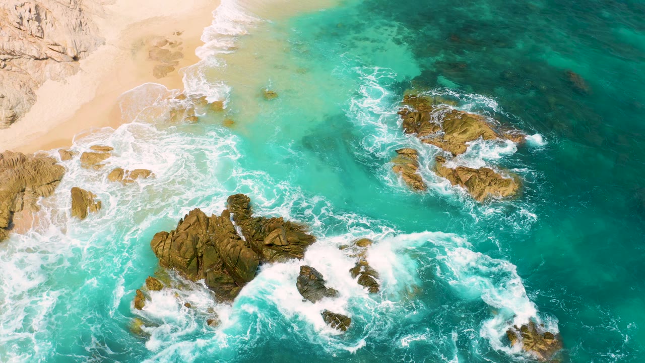 Top-down Mexico coast view with foamy water wrapping around central and right-side rocks, pale beach at the upper left, and no visible people.