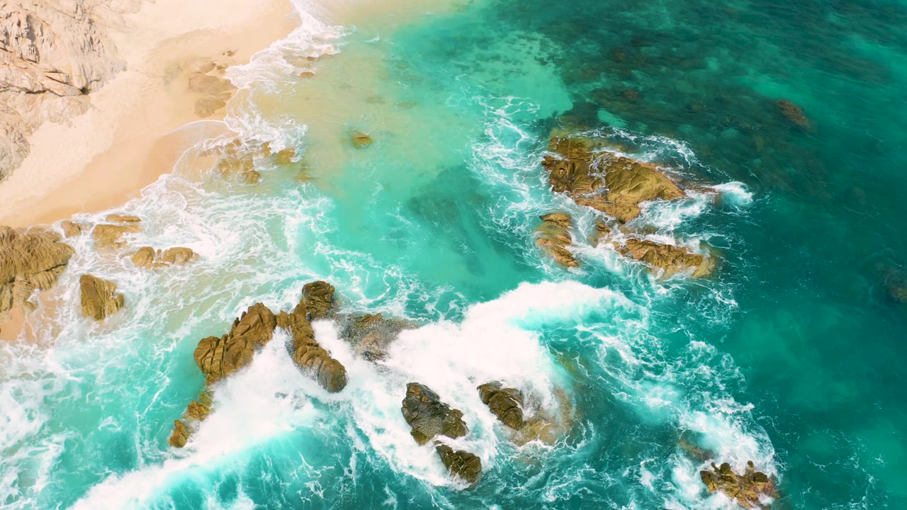 Aerial Mexico shoreline frame with a larger whitewater band crossing the lower rocks, teal water to the right, sand to the upper left, and no visible people.