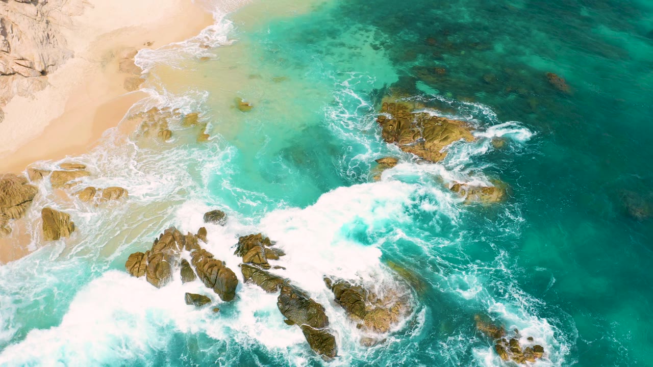 Overhead view of the Mexico coast with white foam spreading from the lower rocks toward the middle, shallow aqua water near the sand, and no visible people.
