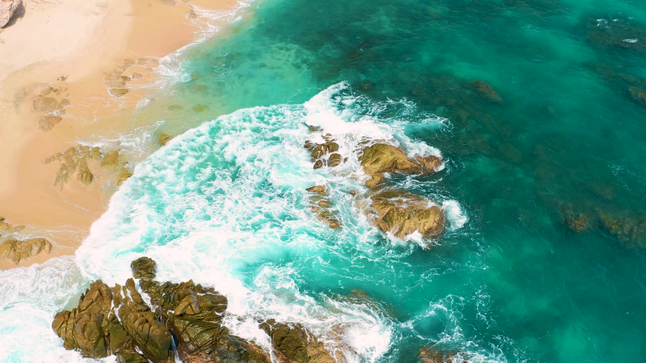 Overhead view of turquoise surf breaking over rocks beside tan beach.