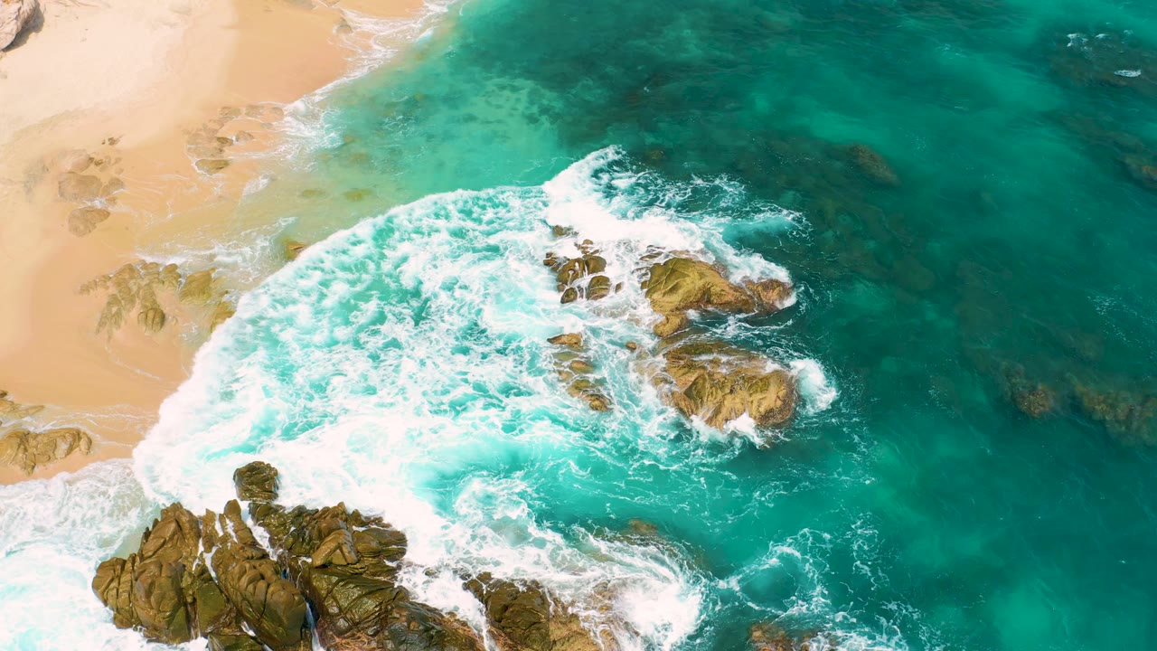 Aerial view at Secrets Marquis Los Cabos, Mexico with white foam expanding around exposed rocks and a sandy beach along the upper left, with no visible people.