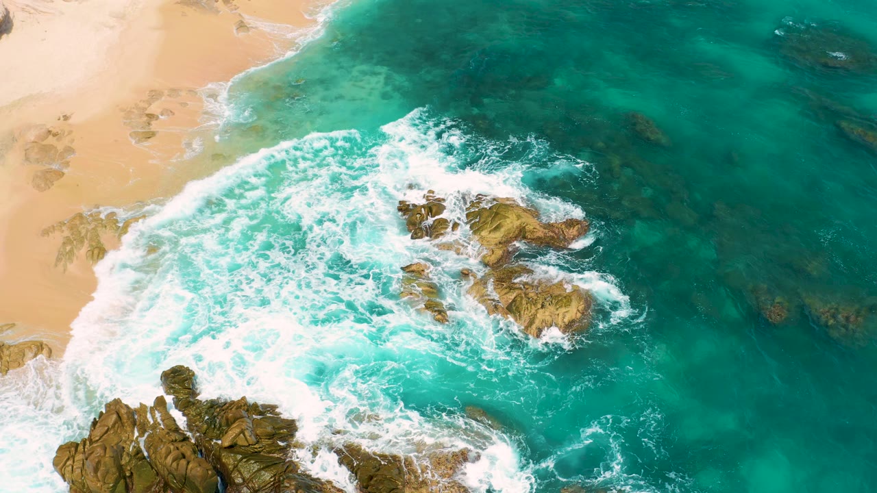 Overhead coastal view from Secrets Marquis Los Cabos, Mexico showing foamy surf curling around central rock outcrops with clear green-blue water to the right, with no visible people.
