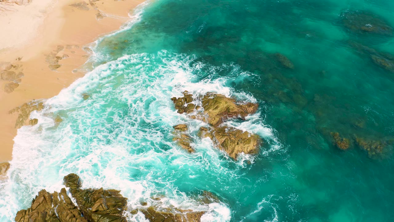 Drone-like overhead frame at Secrets Marquis Los Cabos, Mexico with surf pushing diagonally across clustered rocks, beach on the left, and darker ocean water to the right, with no visible people.