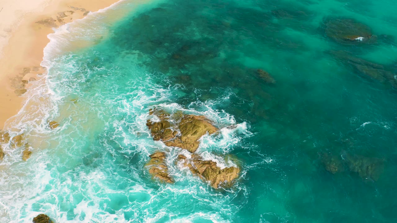 Overhead frame at Secrets Marquis Los Cabos, Mexico with the sandy shoreline along the left, green-blue water across the frame, foam curling around midframe rocks, and no visible people.