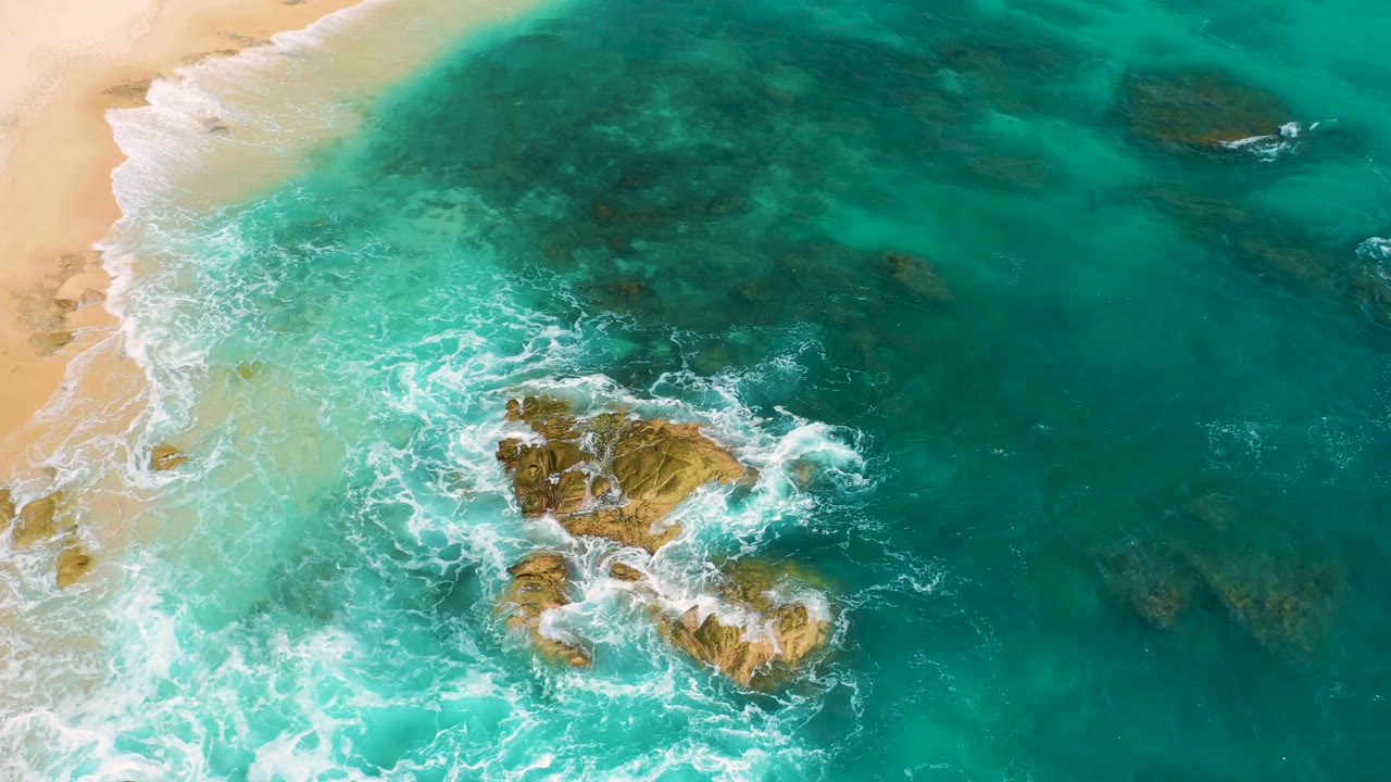 Aerial frame at Secrets Marquis Los Cabos, Mexico showing a wider spread of clear teal water, pale beach at the upper left, two tan rock outcrops, active white foam, and no visible people.