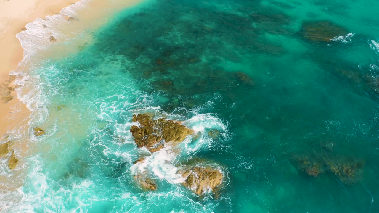 Drone-style aerial frame at Secrets Marquis Los Cabos, Mexico with frothy surf spreading through shallow turquoise water, beach visible along the left side, rocks lower center, and no visible people.