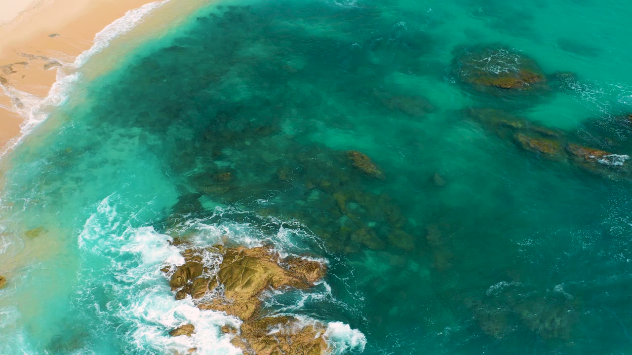 Overhead view at Secrets Marquis Los Cabos, Mexico, with pale sand at the upper left, turquoise water over dark submerged rocks, white surf around exposed rocks, and no visible people.