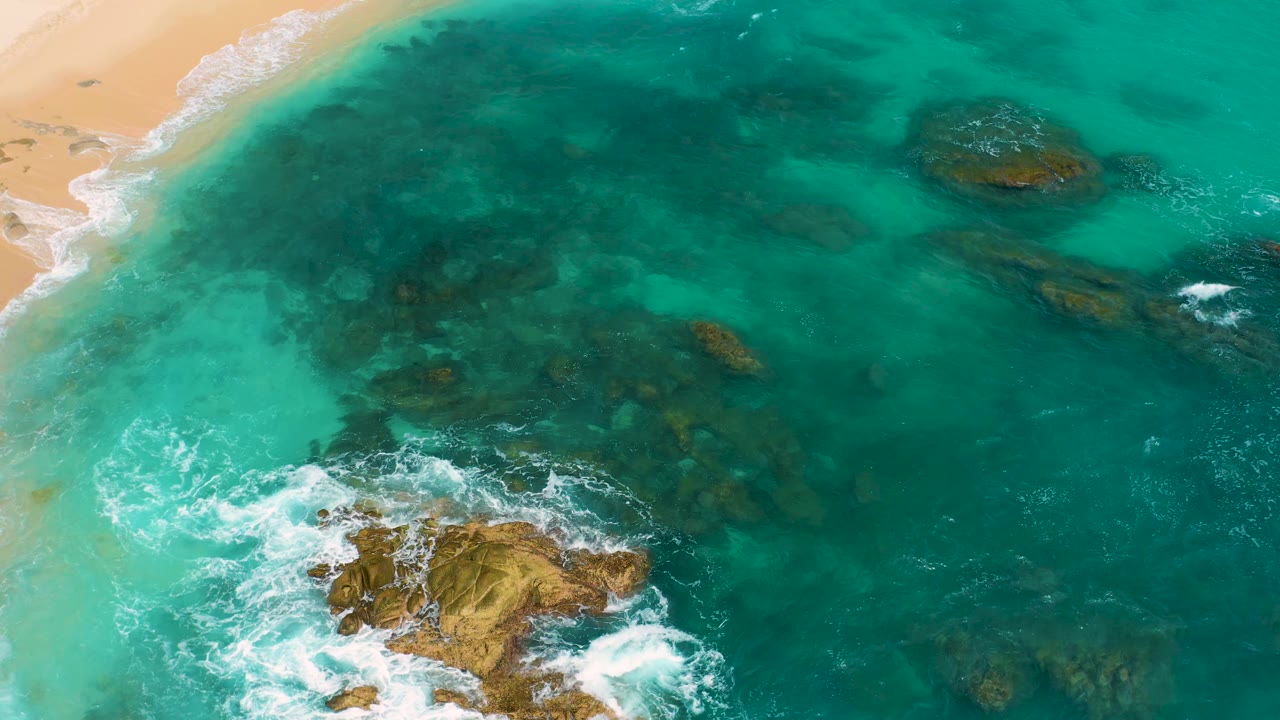 Aerial view at Secrets Marquis Los Cabos, Mexico, with the sandy beach clipped along the upper left, broad turquoise water, brown rock outcrops, white surf at the lower center, and no visible people.