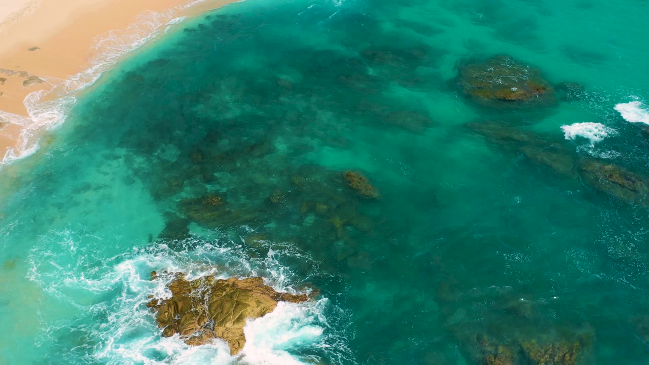 Overhead shot at Secrets Marquis Los Cabos, Mexico, with sand along the upper left, green-blue water filling most of the frame, whitewater around lower rock clusters, and no visible people.