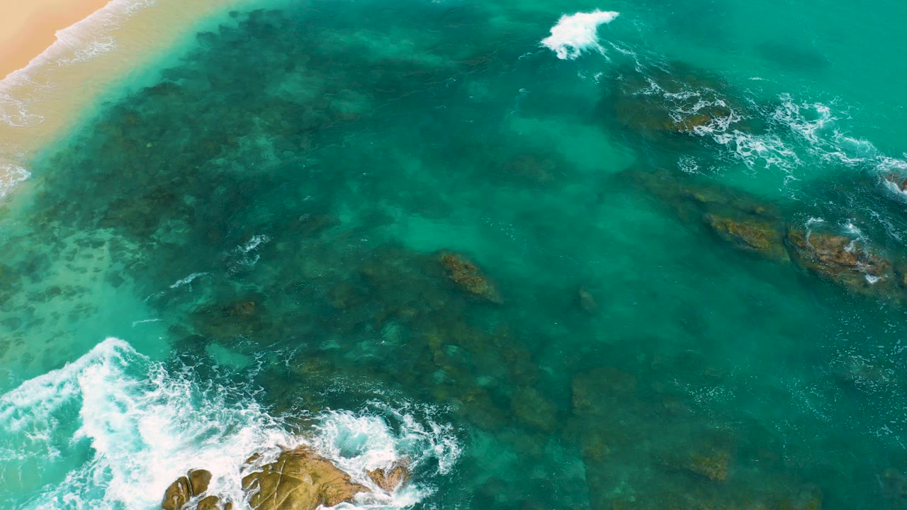 Overhead turquoise ocean with surf, rocks, and a strip of sand.