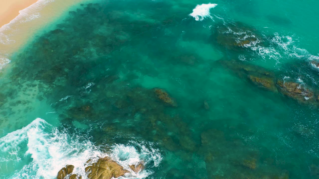 Aerial frame from Secrets Marquis Los Cabos, Mexico, with green-blue water, exposed rocks at the lower edge, white foam moving through the shallows, and no visible people.