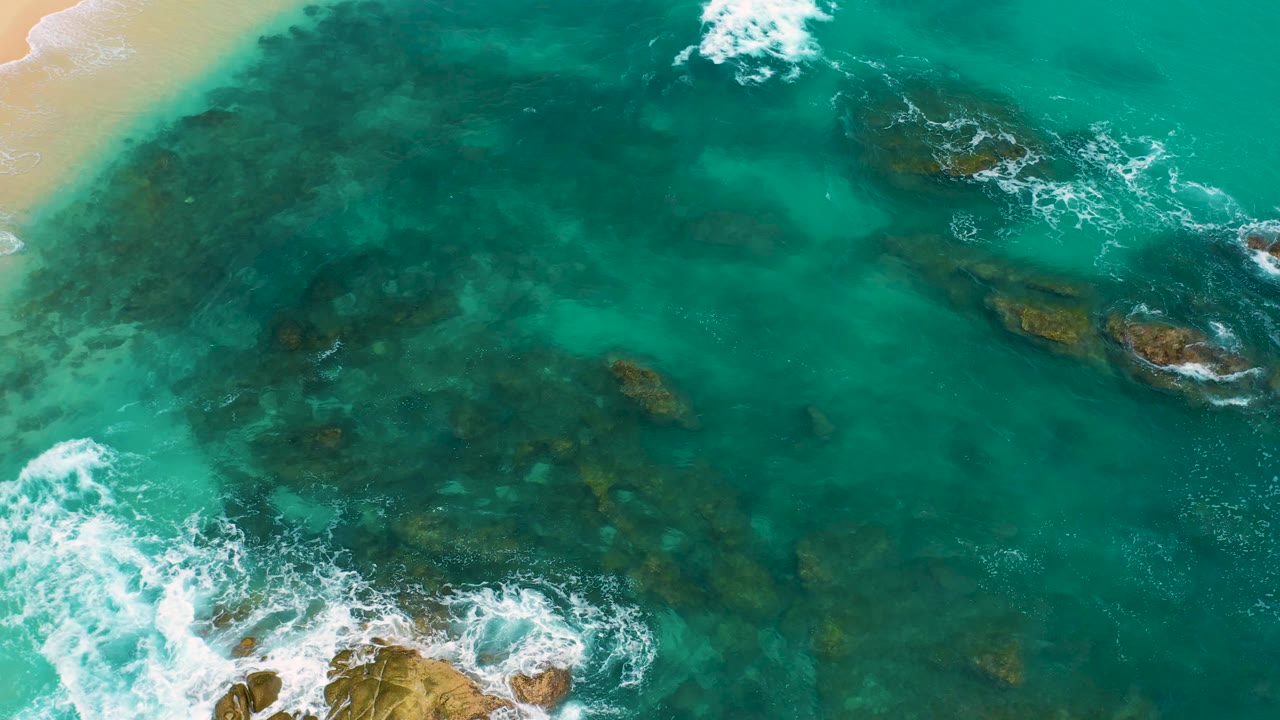 Top-down coastal view at Secrets Marquis Los Cabos, Mexico, with darker submerged rock patches under clear green water, white wave crests near the top and lower left, and no visible people.