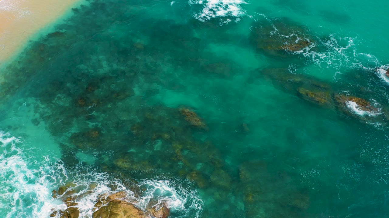 Aerial view over Secrets Marquis Los Cabos, Mexico, where turquoise shallows cover scattered rocks, white foam enters from the lower left, and no visible people appear on the beach or water.