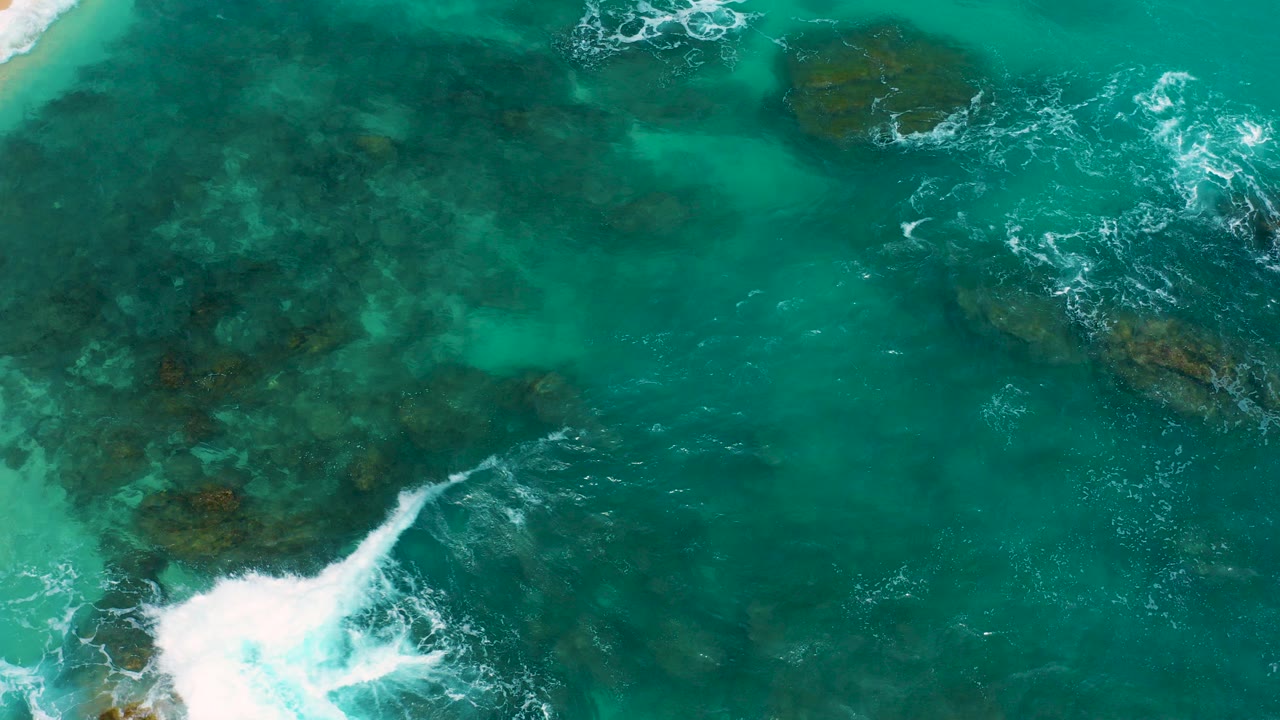 Overhead view at Secrets Marquis Los Cabos, Mexico, with turquoise water covering dark submerged rocks and white surf entering at the lower left, no visible people.