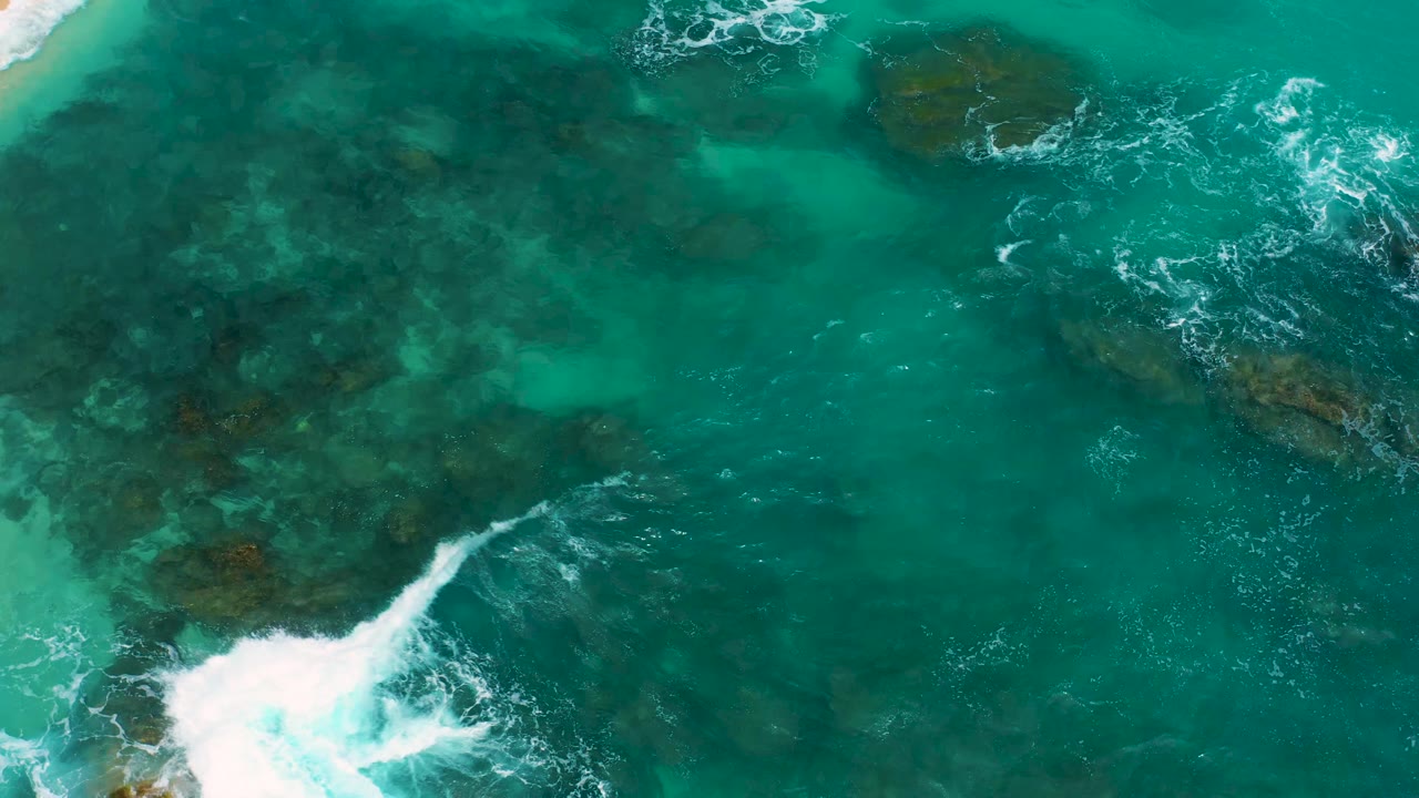 Overhead coastal frame from Secrets Marquis Los Cabos, Mexico, showing white foam spreading across green-blue water above scattered dark rocks, no visible people.